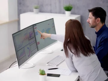 Man and women looking at dual computer screens on white desk in office pointing to website data