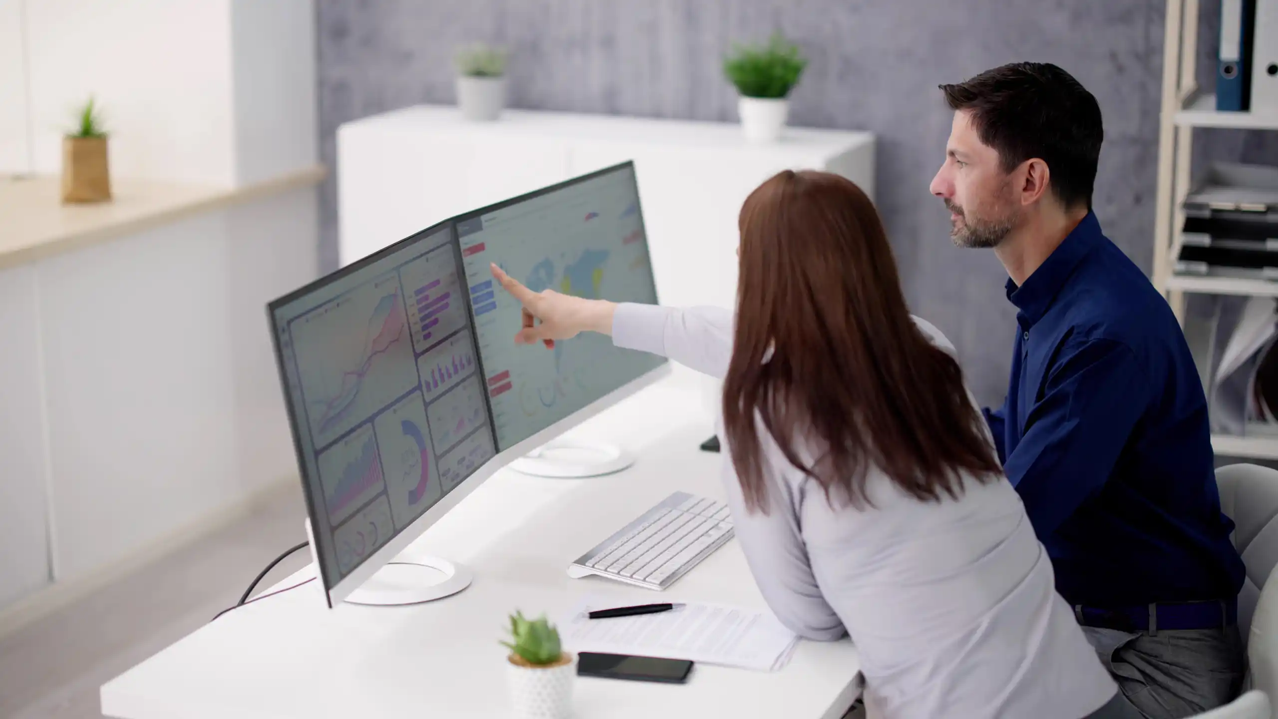 Man and women looking at dual computer screens on white desk in office pointing to website data