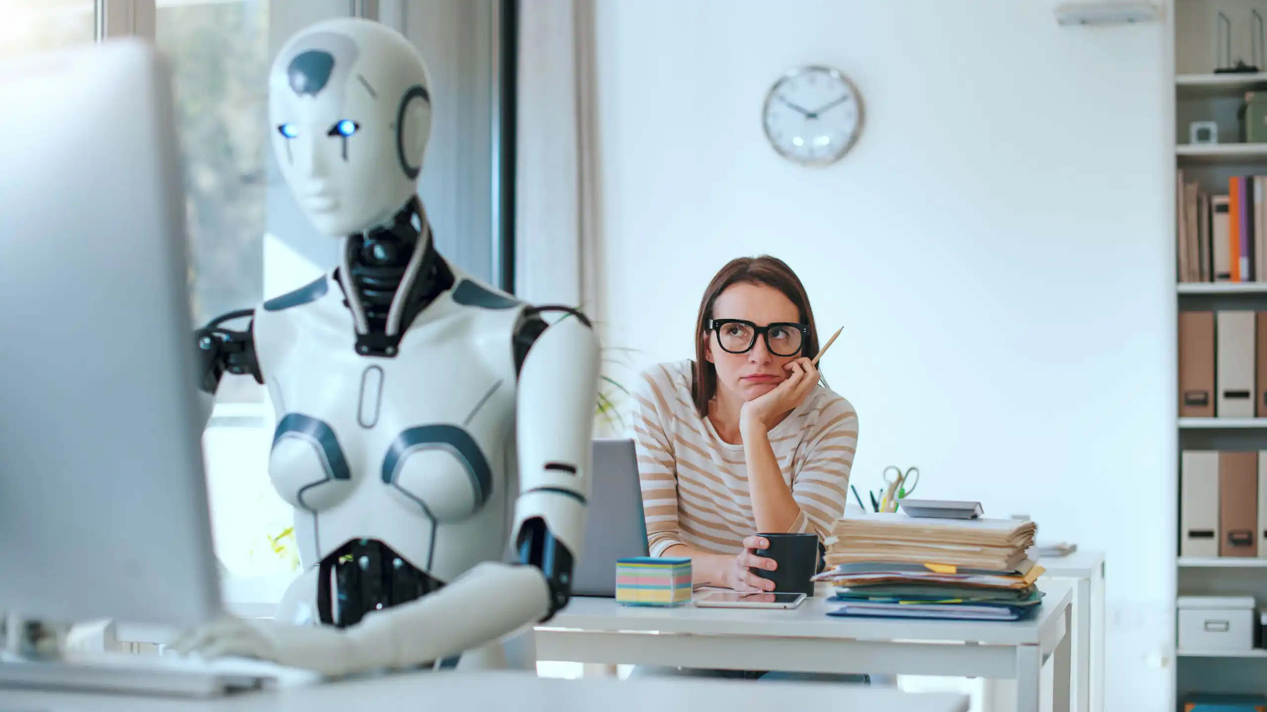 AI agent sitting at computer desk with woman staring at her from behind.